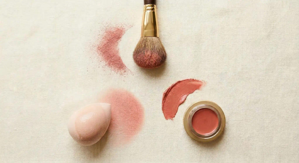 Triangular arrangement of a fluffy blush brush, a damp beauty sponge, and a hand with a cream blush swatch on the fingertip on a warm cream surface