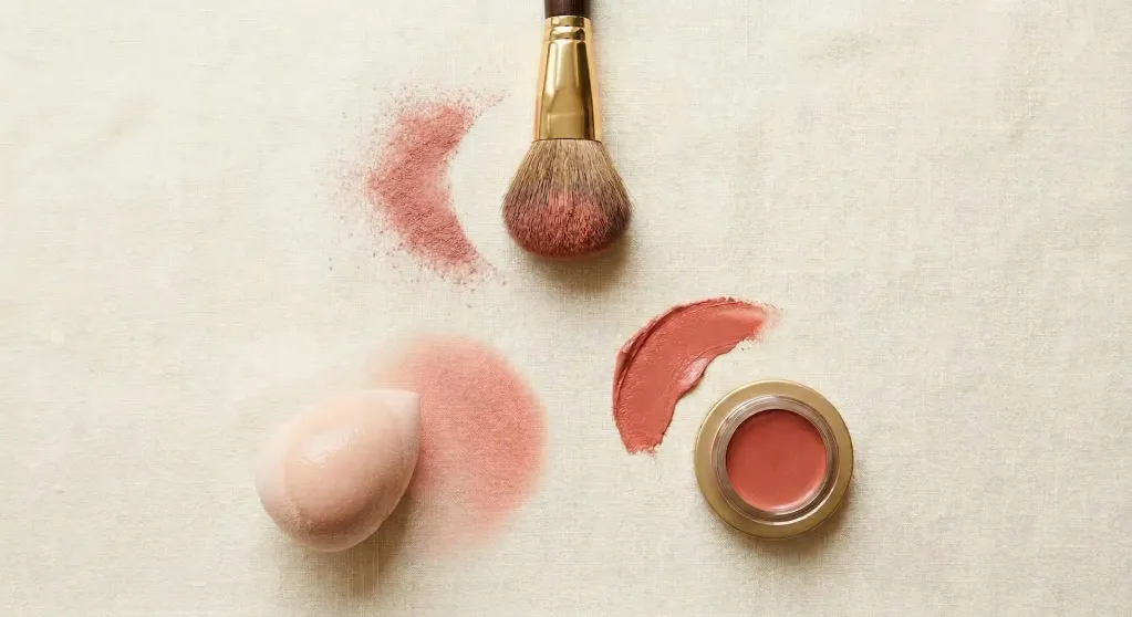 Triangular arrangement of a fluffy blush brush, a damp beauty sponge, and a hand with a cream blush swatch on the fingertip on a warm cream surface