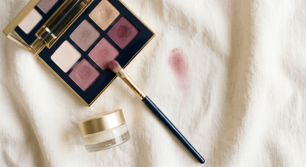 A neutral eyeshadow palette beside a clear lip balm and a small flat brush on a cream surface, with a muted rose pigment swatch