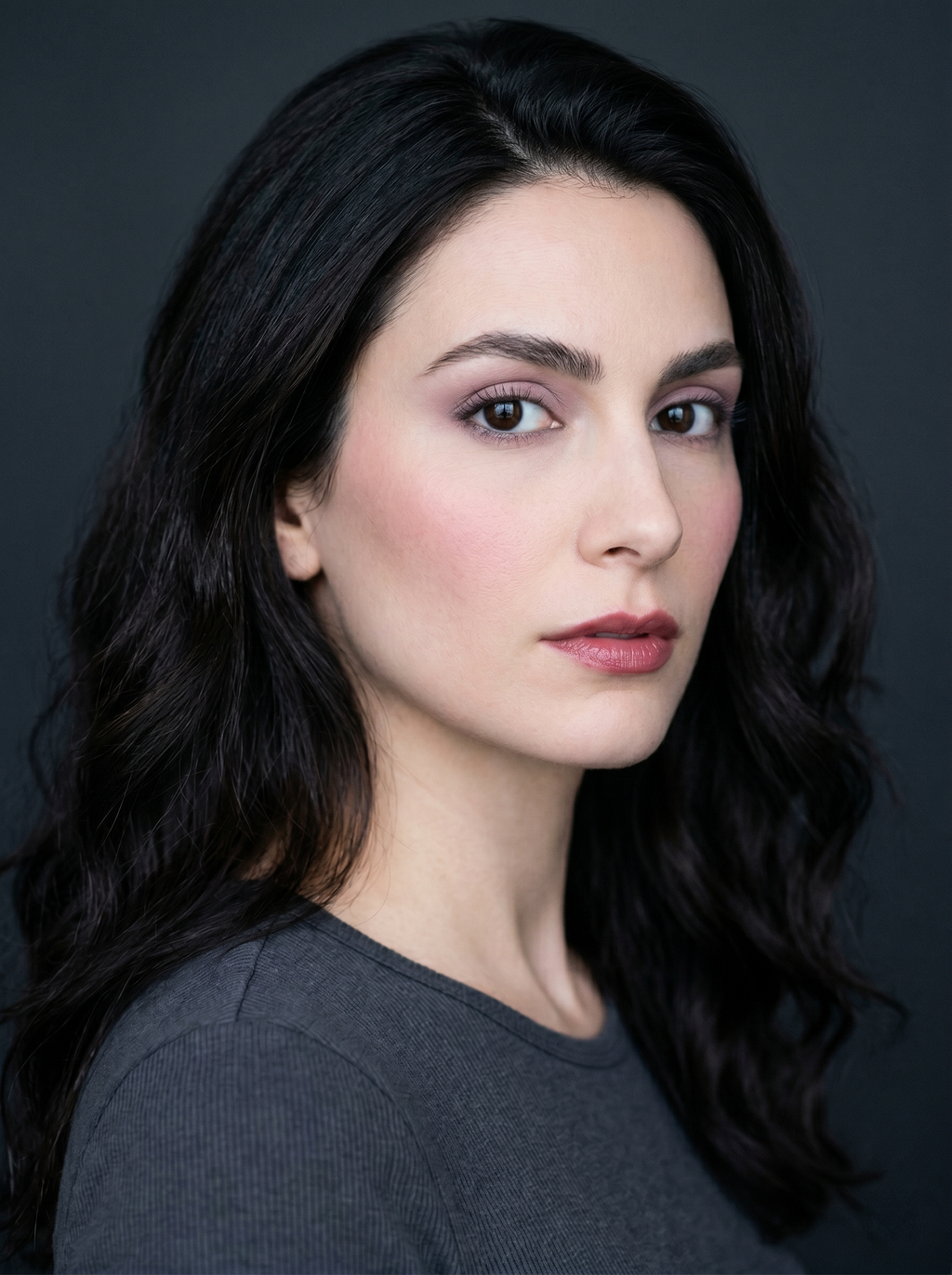 Dark Winter clean girl makeup on fair cool-toned skin: a soft mauve wash on the lids, cool rose blush, groomed dark brows, and a cool berry-nude lip against a charcoal background