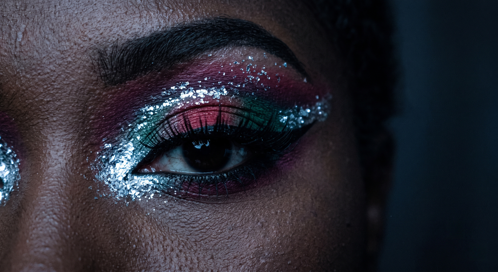Close-up of a single Dark Winter festival eye with a jet black graphic wing, a cool jewel-toned gradient of red, plum, and emerald, and an icy chrome-silver spark at the inner corner, shot with dramatic single-source lighting against a near-black backdrop